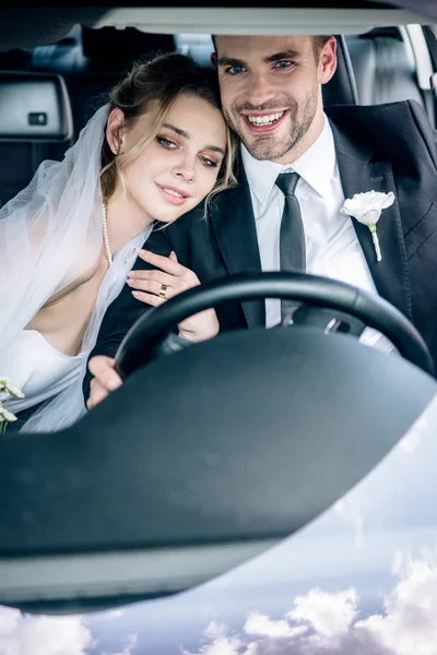 Attractive bride in bridal veil hugging handsome bridegroom in car — Stock Photo