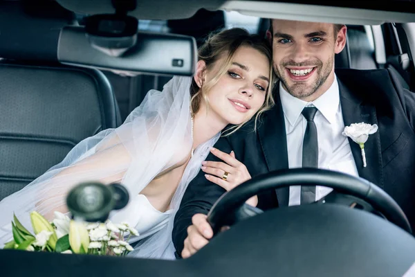 Atractiva novia en el velo nupcial abrazando apuesto novio en coche - foto de stock