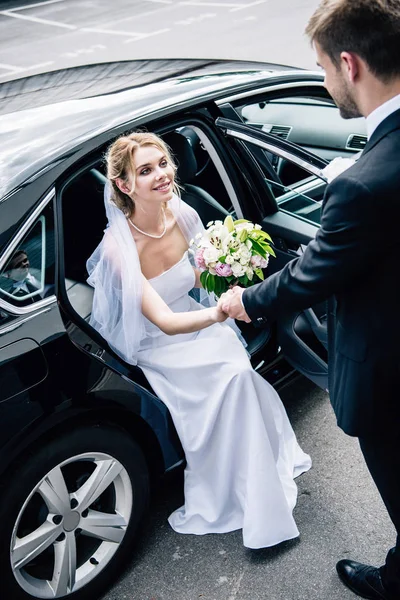 Marié en costume donnant la main à la mariée attrayante et souriante avec bouquet — Photo de stock