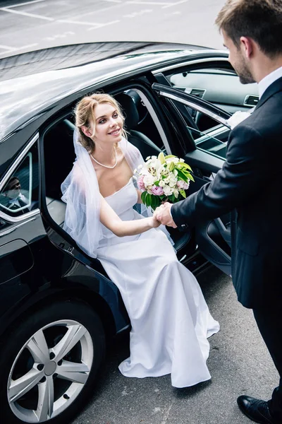 Novio en traje dando la mano a la novia atractiva y sonriente con ramo - foto de stock