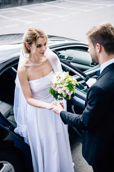 Novio en traje dando la mano a la novia atractiva y sonriente con ramo - foto de stock