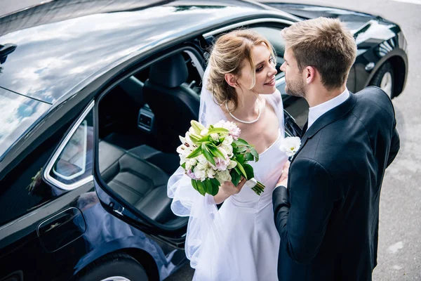 Marié en costume embrasser mariée attrayante et blonde avec bouquet — Photo de stock