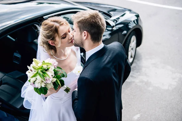 Noivo no terno beijando noiva atraente e loira com buquê — Fotografia de Stock