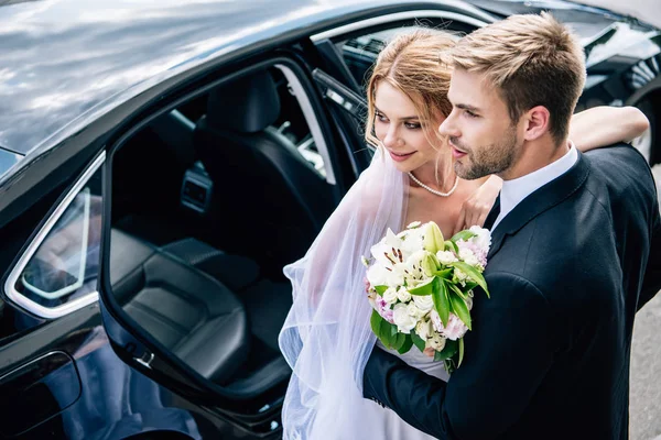 Handsome bridegroom in suit hugging attractive and blonde bride with bouquet — Stock Photo