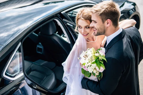 Handsome bridegroom in suit hugging attractive and blonde bride with bouquet — Stock Photo