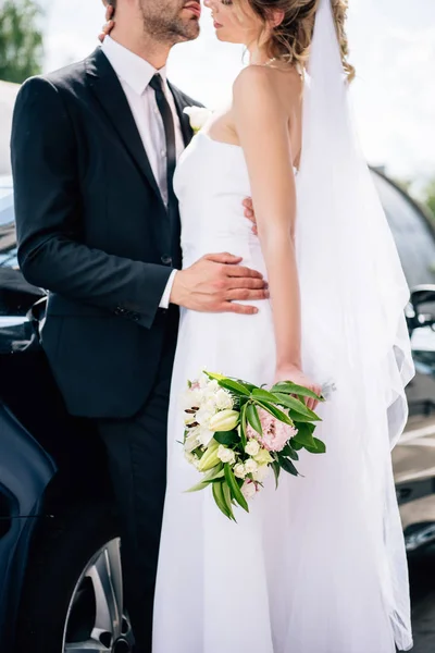 Vista cortada do noivo em terno abraçando noiva no vestido de noiva com buquê — Fotografia de Stock