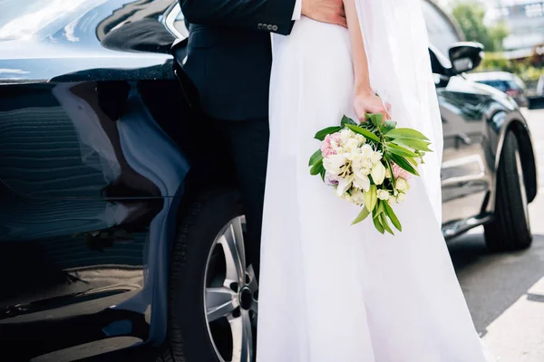 Vista recortada del novio en traje abrazando novia en vestido de novia con ramo - foto de stock