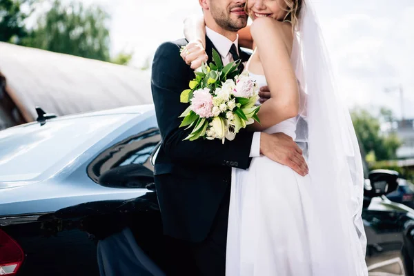 Abgeschnittene Ansicht des Bräutigams im Anzug umarmt Braut im Brautkleid mit Strauß — Stockfoto