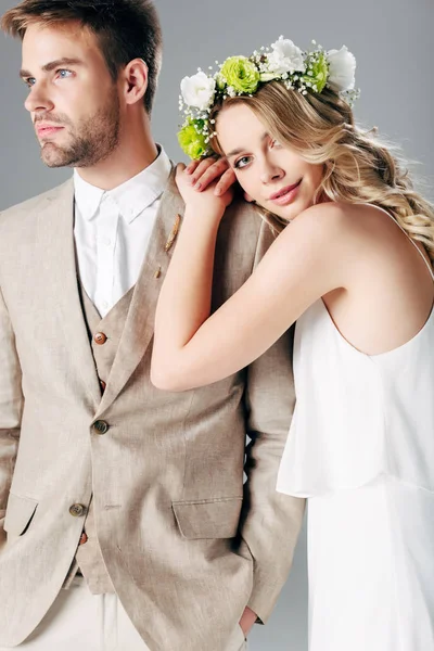 Bride in wedding dress and wreath hugging handsome bridegroom in suit isolated on grey — Stock Photo