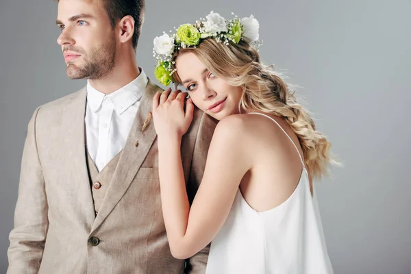 Bride in wedding dress and wreath hugging handsome bridegroom in suit isolated on grey — Stock Photo