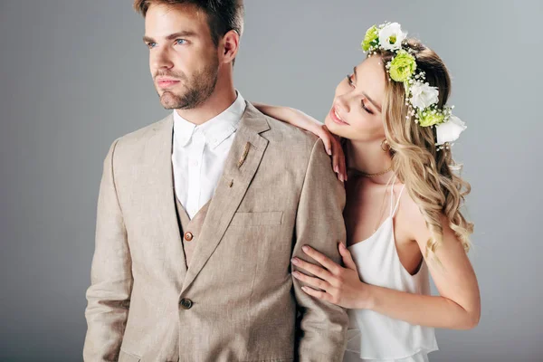 Braut in Brautkleid und Kranz umarmt gutaussehenden Bräutigam im Anzug isoliert auf grau — Stockfoto