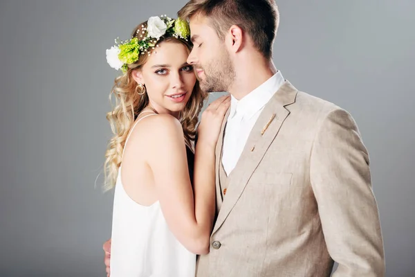 Schöner Bräutigam im Anzug umarmt Braut in Brautkleid und Kranz isoliert auf grau — Stockfoto