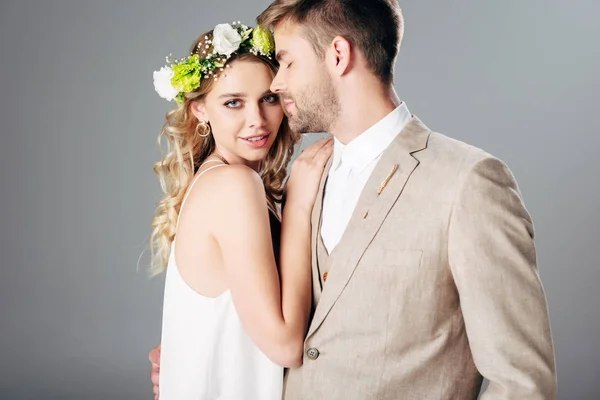 Handsome bridegroom in suit hugging with bride in wedding dress and wreath isolated on grey — Stock Photo