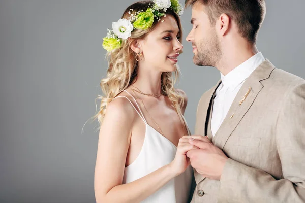 Handsome bridegroom in suit hugging with bride in wedding dress and wreath isolated on grey — Stock Photo