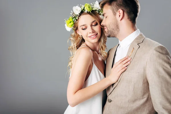 Handsome bridegroom in suit hugging with bride in wedding dress and wreath isolated on grey — Stock Photo