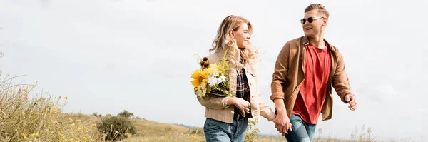 Plano panorámico de mujer atractiva con ramo y hombre guapo sonriendo y cogido de la mano - foto de stock