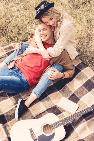 Vista de ángulo alto del hombre y la mujer guapos abrazando y sentado en la manta - foto de stock