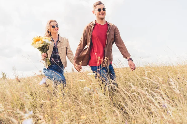 Atractiva mujer y hombre guapo en gafas de sol caminando y tomados de la mano - foto de stock