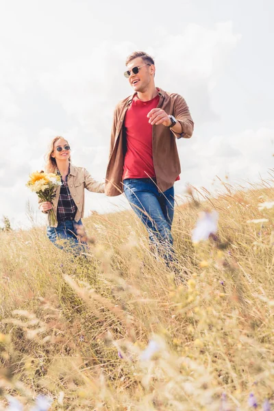 Atractiva mujer y hombre guapo en gafas de sol caminando y tomados de la mano - foto de stock