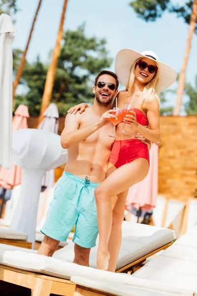 Ganzkörperansicht des glücklichen lächelnden Paares, das Cocktails hält und sich bei sonnigem Tag im Resort umarmt — Stockfoto