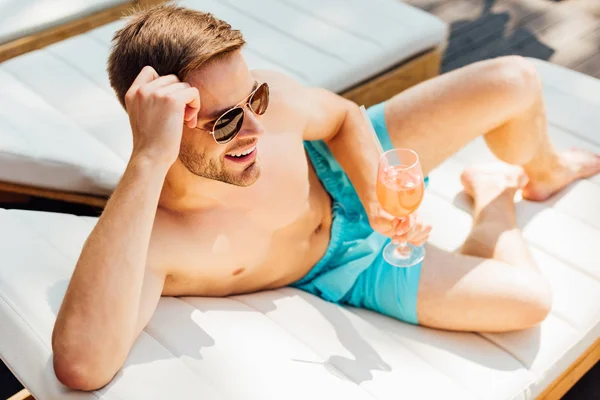 Happy shirtless man in sunglasses lying on lounger and holding glass of cocktail at resort — Stock Photo