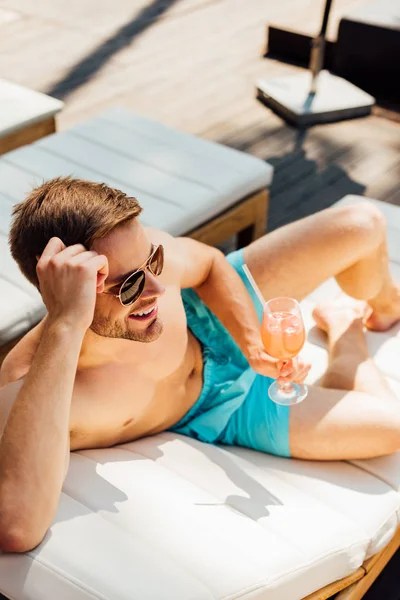 Hombre feliz sin camisa en gafas de sol acostado en una tumbona y sosteniendo un vaso de cóctel en el resort - foto de stock