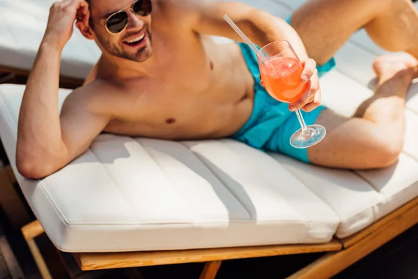 Счастливый мужчина без рубашки в солнечных очках лежит на шезлонге и держит стакан коктейля на курорте — стоковое фото