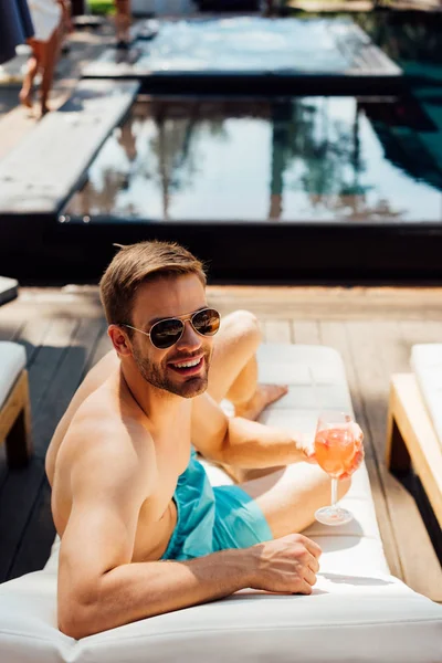 Felice uomo senza maglietta in occhiali da sole sdraiato sul lettino e con in mano un bicchiere di cocktail al resort — Foto stock