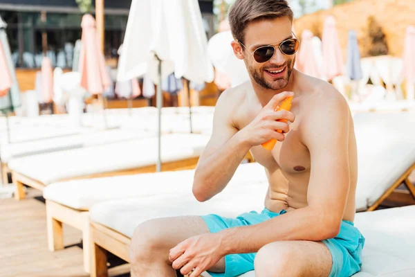 Homem sem camisa sentado na espreguiçadeira e aplicando protetor solar no resort — Fotografia de Stock