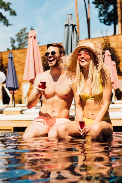 Sexy pareja sosteniendo vasos de vino tinto y sonriendo en la piscina - foto de stock