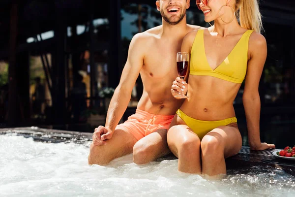 Vista recortada de sexy pareja sonriente con vino tinto y fresas en la piscina - foto de stock