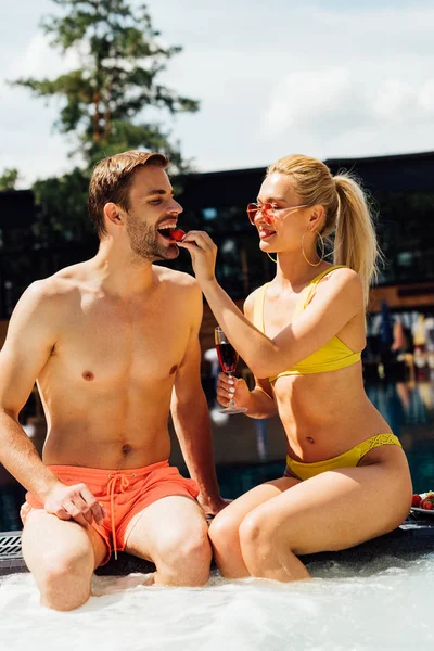 Sexy girl holding glass of red wine and feeding boyfriend with strawberry in swimming pool — Stock Photo