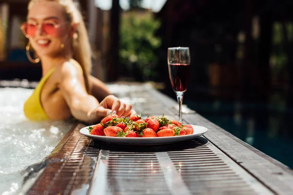 Selective focus of sexy girl in swimsuit with red wine and strawberries in swimming pool — Stock Photo