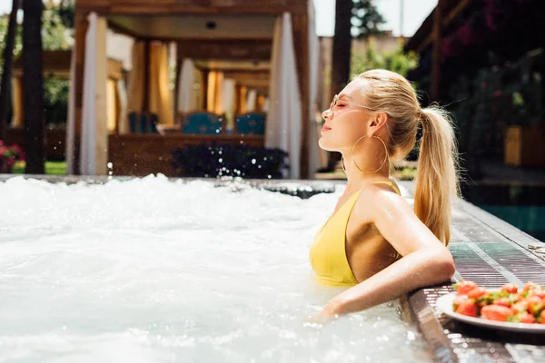 Vue latérale de fille sexy dans des lunettes de soleil avec des fraises dans la piscine — Photo de stock