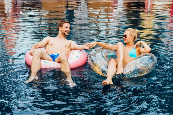 Lächelndes Paar, Händchen haltend und einander anschauend, während sie auf Schwimmringen im Pool liegen — Stockfoto