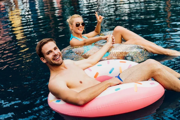 Couple souriant tenant la main tout en étant couché sur des anneaux de natation dans la piscine — Photo de stock