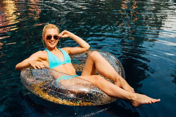 Chica sexy en traje de baño y gafas de sol sonriendo mientras está acostado en el anillo de natación en la piscina - foto de stock