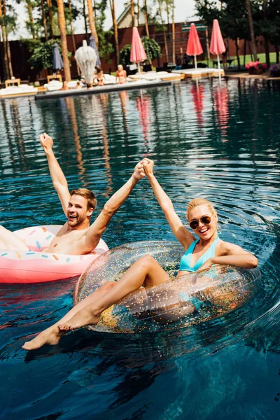 Happy couple holding hands while lying on swim rings in swimming pool — Stock Photo