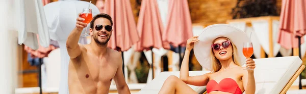 Панорамный снимок улыбающейся пары, отдыхающей на шезлонгах и поднимающей бокалы с коктейлями на курорте — стоковое фото