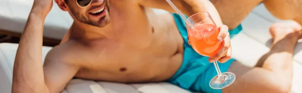 Panoramic shot of laughing shirtless man lying on lounger and holding cocktail — Stock Photo