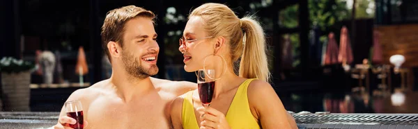 Panoramic shot of sexy couple holding wine glasses with red wine and looking at each other at resort — Stock Photo