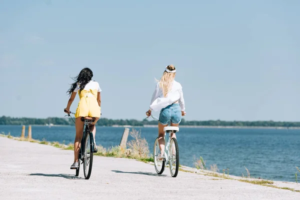 Rückansicht von blonden und brünetten Mädchen, die im Sommer in Flussnähe Fahrrad fahren — Stockfoto