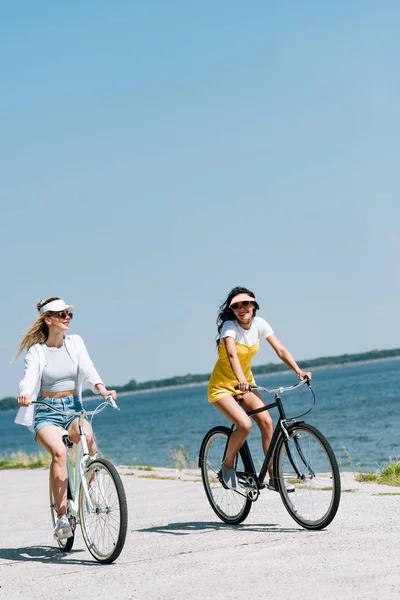 Blonde and brunette girls riding bikes near river in summer — Stock Photo