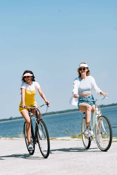 Sorrindo meninas loiras e morenas andando de bicicleta perto do rio no verão — Fotografia de Stock