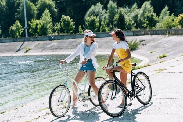 Chicas rubias y morenas felices con bicicletas mirándose el uno al otro cerca del río en verano - foto de stock