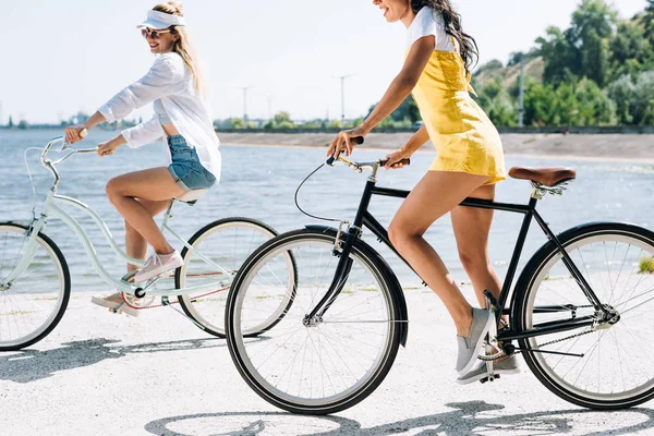Vista lateral de meninas loiras e morenas andando de bicicleta perto do rio no verão — Fotografia de Stock