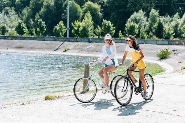Cheerful blonde and brunette girls riding bikes near river in summer — Stock Photo