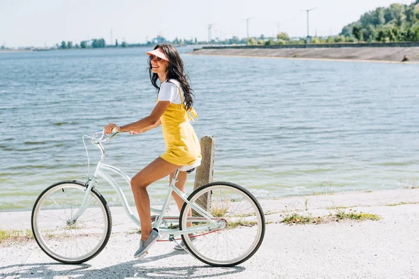 Вид сбоку на счастливую брюнетку красивой девушки, катающейся на велосипеде у реки летом — стоковое фото