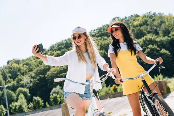 Щаслива блондинка і брюнетка дівчата катаються на велосипедах і приймають селфі влітку — стокове фото