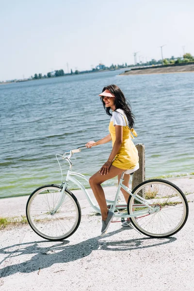 Vue latérale de fille brune heureuse à vélo près de la rivière en été — Photo de stock